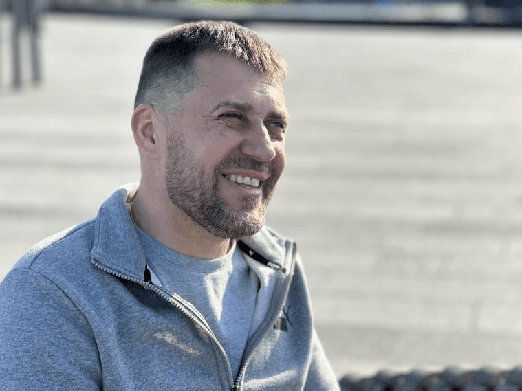 Чоловік із короткою бородою та стрижкою посміхається, сидячи на відкритому повітрі, вдягнений у сіру кофту.