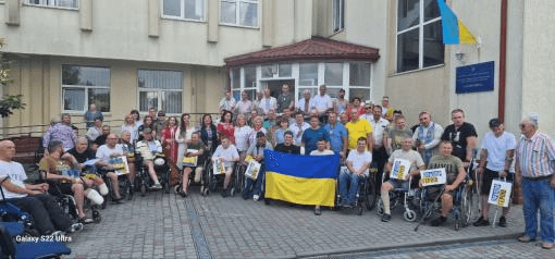 Групове фото людей, серед яких багато в інвалідних візках, перед будівлею з прапором України. У центрі тримають український прапор.