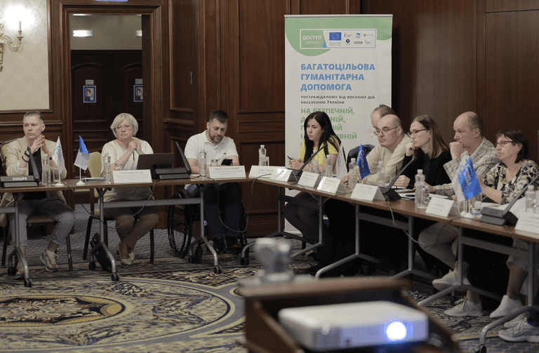 Люди сидять за столом з табличками, на передньому плані проектор лінза якого світиться