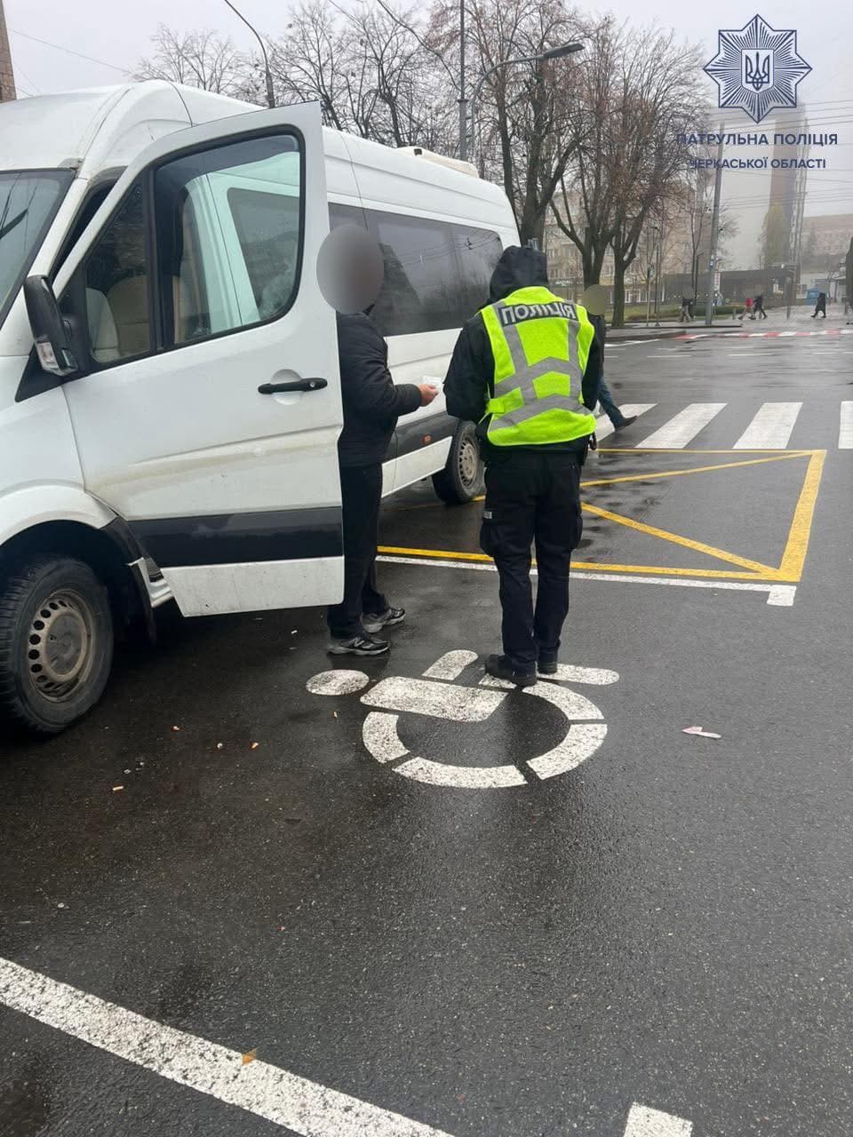 Поліцейський у світловідбивному жилеті з написом «ПОЛІЦІЯ» спілкується з людиною біля білого фургона, припаркованого на місці для людей з інвалідністю. На асфальті видно символ інвалідного візка. Сцена відбувається на мокрій вулиці біля пішохідного переходу, на фоні — будівлі та голі дерева. Зображення може свідчити про перевірку порушення правил паркування. Присутній логотип «Патрульна поліція Черкаської області».